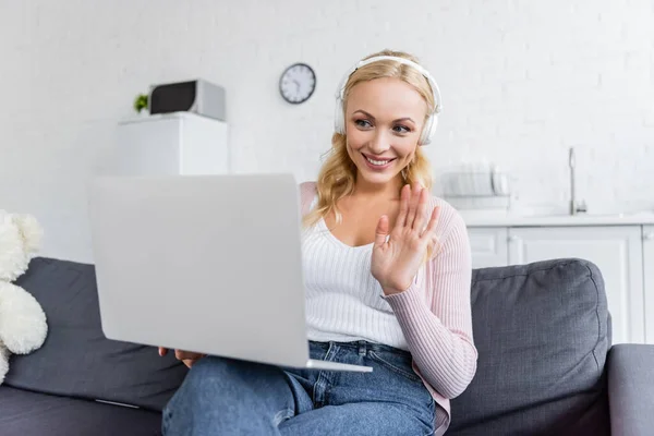 Mulher alegre em fones de ouvido acenando mão durante o bate-papo de vídeo no laptop — Fotografia de Stock