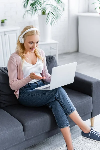 Lächelnde Frau mit drahtlosen Kopfhörern, die während eines Videogesprächs auf ihren Laptop zeigt — Stockfoto