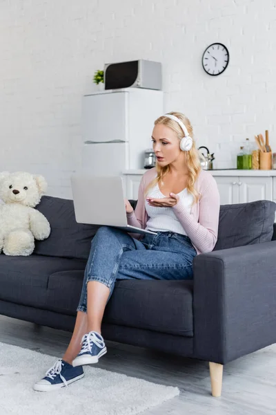 Серьезная женщина в наушниках, указывающая на ноутбук во время видеозвонка на диване рядом с плюшевым мишкой — стоковое фото