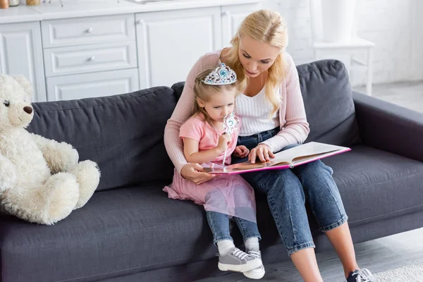 Frau zeigt auf Buch, während sie Tochter in Spielzeugkrone Märchen vorliest — Stockfoto