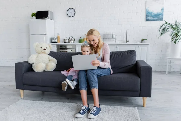 Happy mother and daughter using laptop on couch near teddy bear — Stock Photo