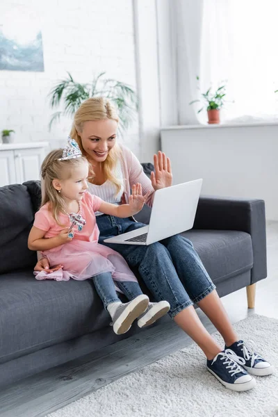 Femme gaie et fille dans la couronne de jouet agitant les mains pendant l'appel vidéo sur ordinateur portable — Photo de stock