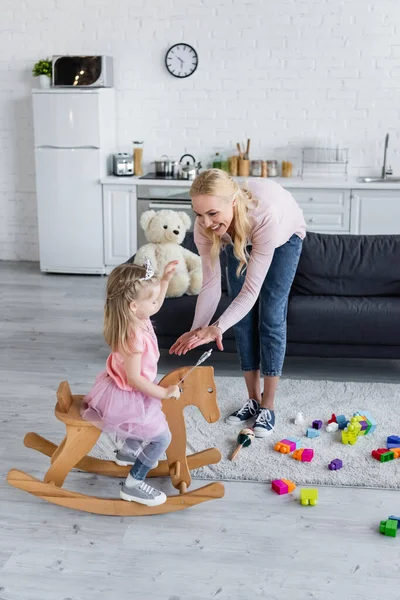Happy woman giving hands to daughter riding rocking horse in costume of princess — Stock Photo
