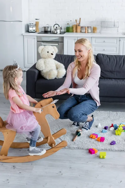 Cheerful mom giving hands to daughter riding rocking horse in fairy costume — Stock Photo