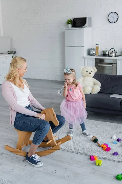 Fröhliche Frau reitet Schaukelpferd neben Tochter im Märchenkostüm — Stockfoto