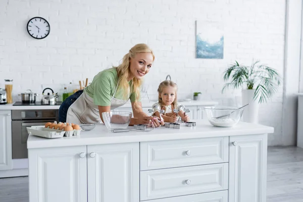 Donna sorridente alla macchina fotografica vicino figlia e tavolo con ingredienti in cucina — Foto stock