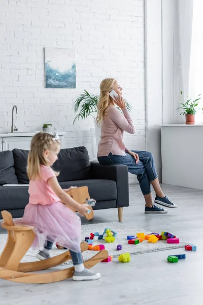 Mulher falando no telefone celular perto de filha borrada montando cavalo de balanço — Fotografia de Stock