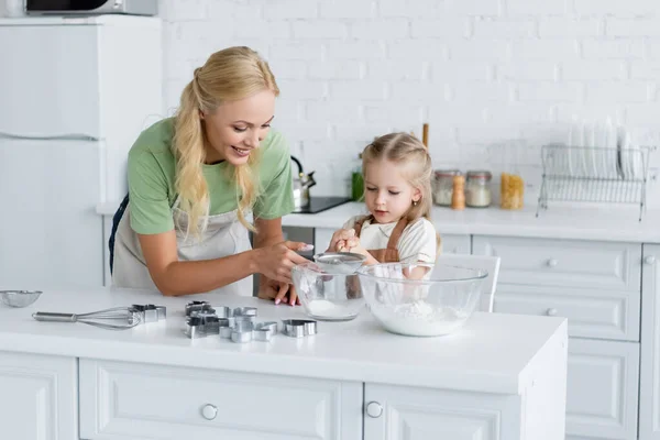 Chica tamizar harina con tamiz en tazón cerca feliz madre - foto de stock