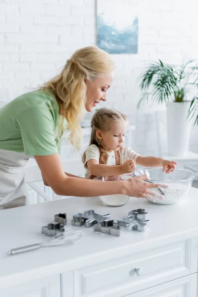 Chica sosteniendo tamiz cerca de tazón con harina mientras ayuda a mamá en la cocina - foto de stock
