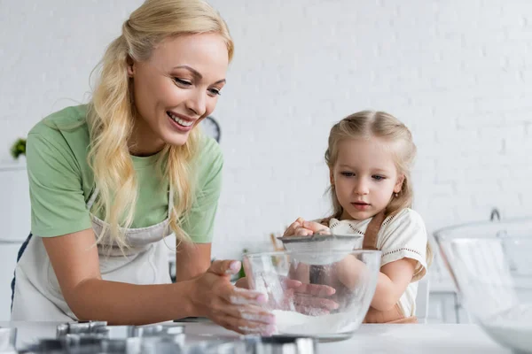 Fröhliche Frau hält Schüssel mit Mehl in der Nähe kleine Tochter mit Sieb — Stockfoto