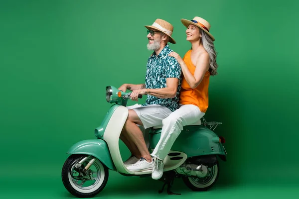 Happy interracial middle aged couple in sun hats riding motor scooter on green — Stock Photo