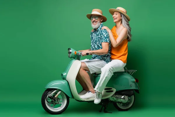 Cheerful interracial middle aged couple in sun hats riding moped on green — Stock Photo