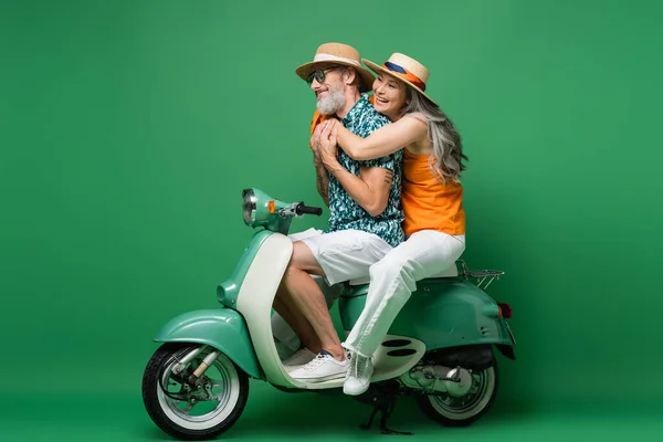 Fröhliche und interrassische Paare mittleren Alters in Sonnenhüten, die sich umarmen, während sie mit dem Moped auf Grün fahren — Stockfoto