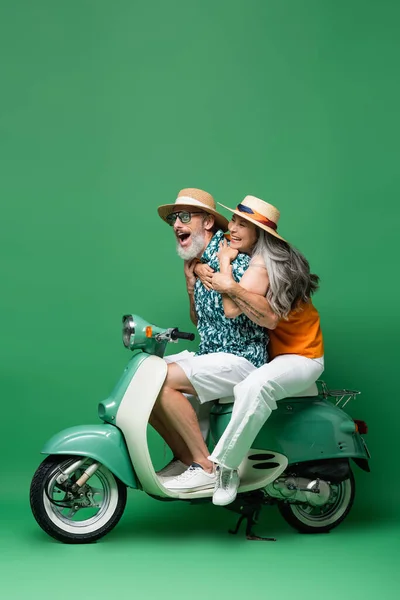 Збуджена і міжрасова пара середнього віку в сонячних капелюхах, обіймаючись під час їзди на моторолері на зеленому — стокове фото