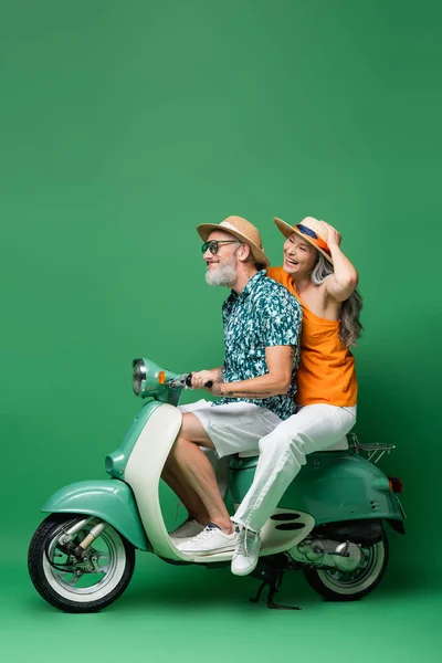 Positivo e interracial casal de meia idade em chapéus de sol andar de scooter motor em verde — Fotografia de Stock