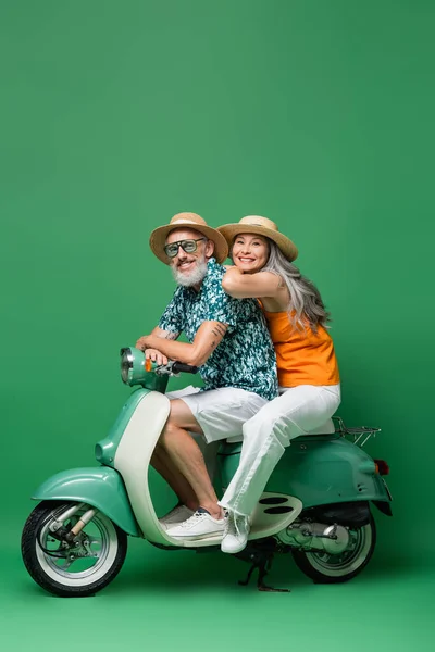 Interracial casal de meia idade em chapéus de palha sorrindo e montando ciclomotor em verde — Fotografia de Stock