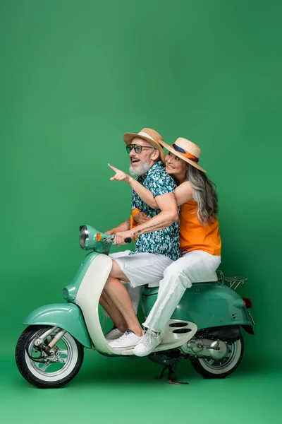 Happy asian middle aged woman pointing with finger and hugging husband while riding motor scooter on green — Stock Photo