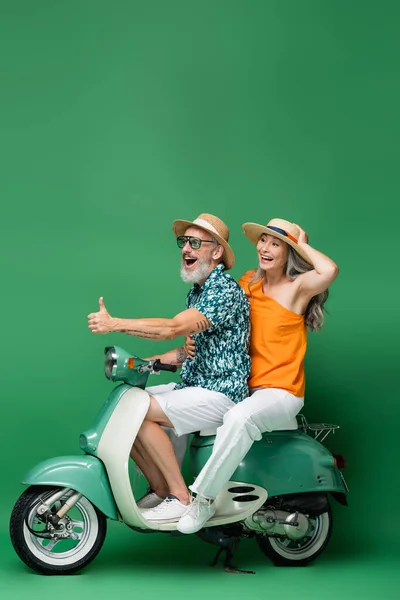 Felice uomo di mezza età mostrando pollice in su mentre cavalca con moglie asiatica su ciclomotore su verde — Foto stock