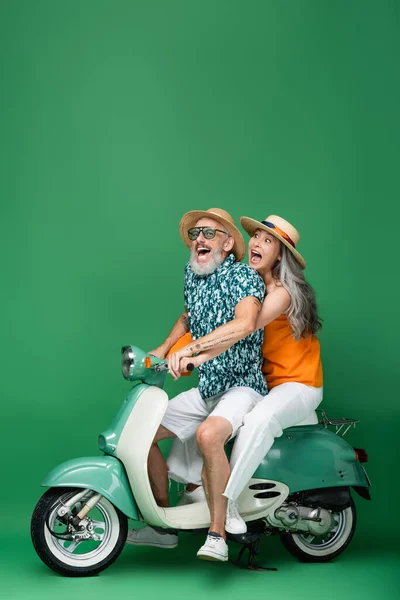 Excited multiethnic and middle aged couple in sun hats riding motor scooter on green — Stock Photo