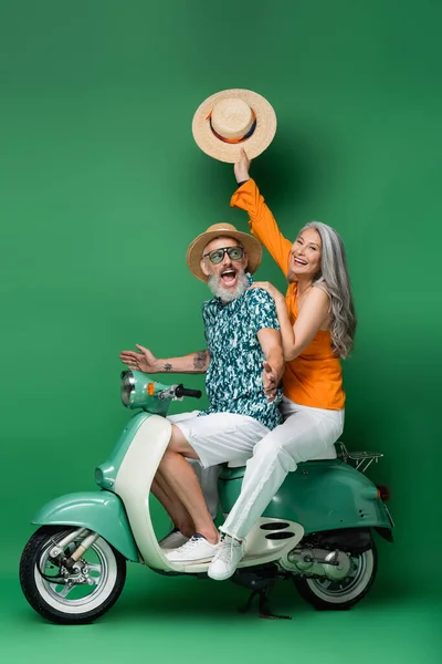 Feliz de mediana edad mujer asiática sosteniendo sombrero de sol mientras monta ciclomotor con excitado marido en verde - foto de stock