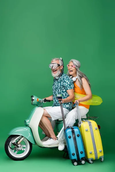 Excited and multiethnic middle aged couple in goggles holding passports and luggage while riding motor scooter on green — Stock Photo