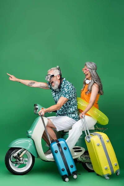 Amazed and middle aged man in goggles pointing with finger near asian wife with luggage while riding motor scooter on green — Stock Photo