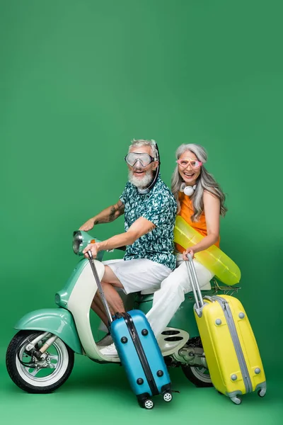 Multiethnique et heureux couple d'âge moyen dans des lunettes tenant des bagages tout en chevauchant cyclomoteur sur vert — Photo de stock