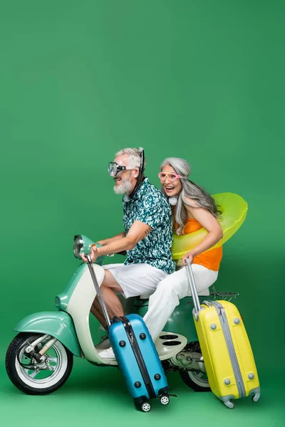Multiethnic and cheerful middle aged couple in goggles holding luggage while riding motor scooter on green — Stock Photo