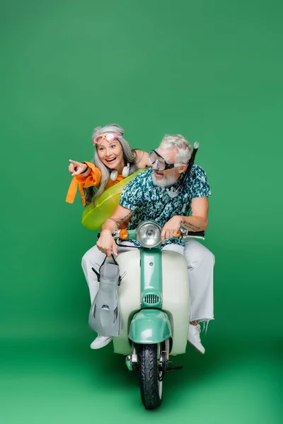 Cheerful middle aged asian woman pointing with finger near husband holding flippers while riding motor scooter on green — Stock Photo