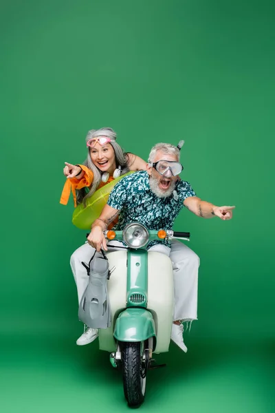 Cheerful and multiethnic middle aged couple in goggles pointing with fingers while riding moped on green — Stock Photo