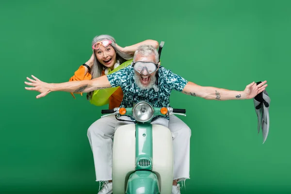 Cheerful middle aged man with outstretched hands holding flippers near asian wife in goggles riding moped on green — Stock Photo