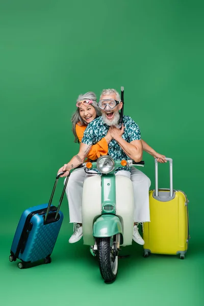 Amazed and mature multiethnic couple in goggles holding luggage while riding moped on green — Stock Photo