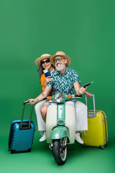 Cheerful middle aged and multiethnic couple in sun hats holding passports and luggage while riding moped on green — Stock Photo