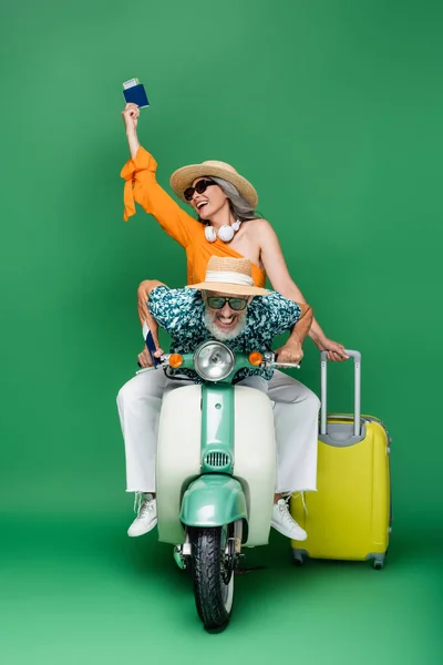 Multiétnico e alegre casal de meia-idade em chapéus de sol segurando passaportes com passagens aéreas enquanto montava ciclomotor em verde — Fotografia de Stock