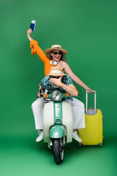 Веселая азиатка средних лет в шляпе на солнце держит паспорт рядом с мужем прикрывая глаза во время езды на мотороллере на зеленом — стоковое фото
