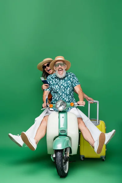 Interracial e positivo casal de meia idade em chapéus de sol segurando passaportes com passagens aéreas enquanto montava ciclomotor em verde — Fotografia de Stock