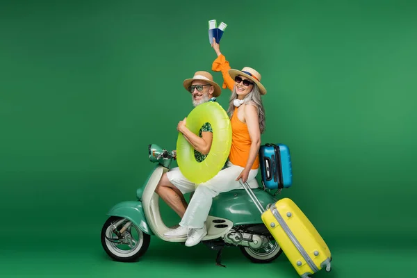 Fröhliche asiatische Frau mittleren Alters mit Sonnenhut hält Pässe in der Nähe Ehemann hält Schwimmring, während Moped fahren auf grün — Stockfoto