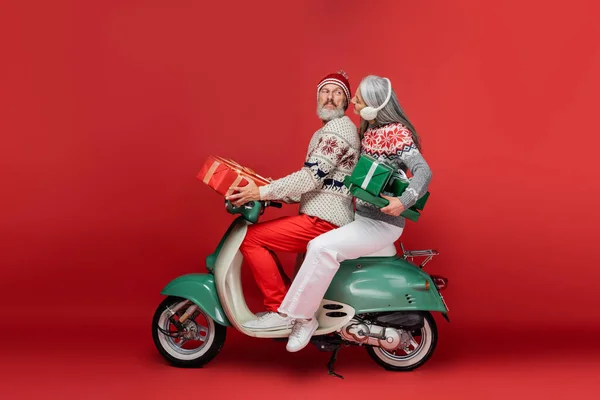 Cheerful middle aged and asian woman in ear muffs holding christmas presents while riding moped with husband on red — Stock Photo