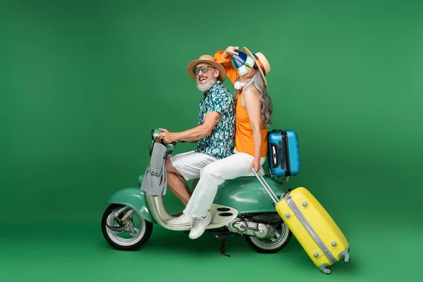 Доросла азійка у сонцезахисному капелюсі закриває обличчя паспортами біля чоловіка, тримаючи ласти під час їзди мопедом на зеленому. — стокове фото