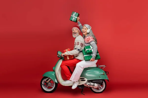 Cheerful middle aged asian woman in ear muffs holding christmas presents while riding motor scooter with excited husband on red — Stock Photo