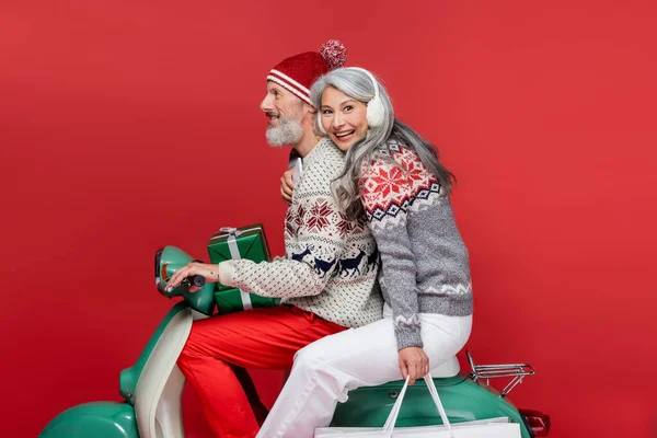 Happy and interracial mature couple with christmas presents riding moped on red — Stock Photo