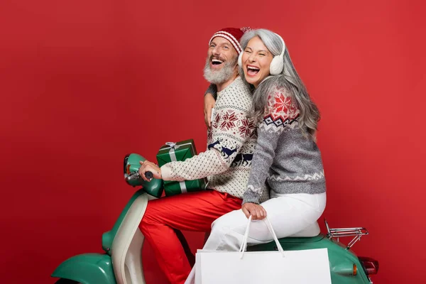 Excited and interracial mature couple with christmas presents riding moped on red — Stock Photo