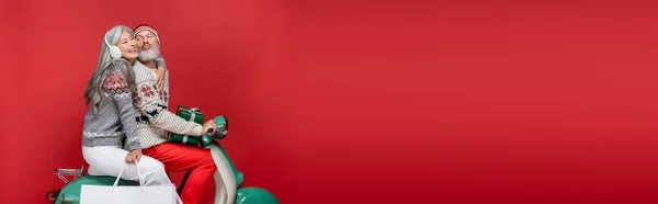 Homme d'âge moyen avec Noël présente des lèvres boudantes près de la femme asiatique tout en chevauchant scooter moteur sur rouge, bannière — Photo de stock