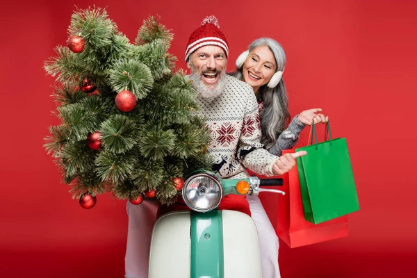 Cheerful middle aged man holding christmas tree near asian wife while riding moped on red — Stock Photo