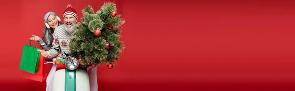 Eccitato uomo di mezza età che tiene l'albero di Natale vicino alla moglie asiatica mentre cavalca il motorino sul rosso, banner — Foto stock