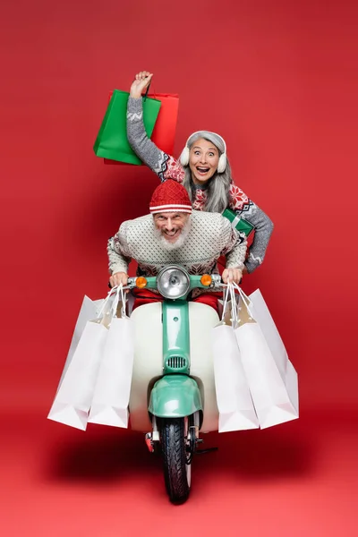 Animado meia idade interracial casal em camisolas com compras sacos equitação ciclomotor no vermelho — Fotografia de Stock