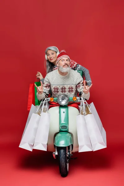 Hombre de mediana edad mostrando señal de roca cerca sorprendido esposa asiática con bolsas de compras mientras monta moto en rojo - foto de stock