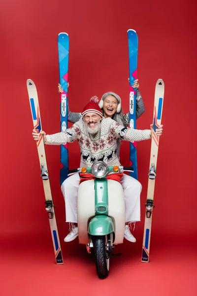 Happy interracial middle aged couple in sweaters holding skis while riding moped on red — Stock Photo
