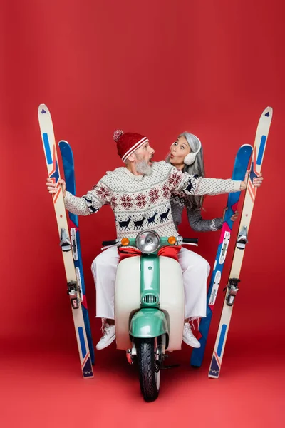 Surprised and interracial middle aged couple in sweaters holding skis while riding motor scooter on red — Stock Photo