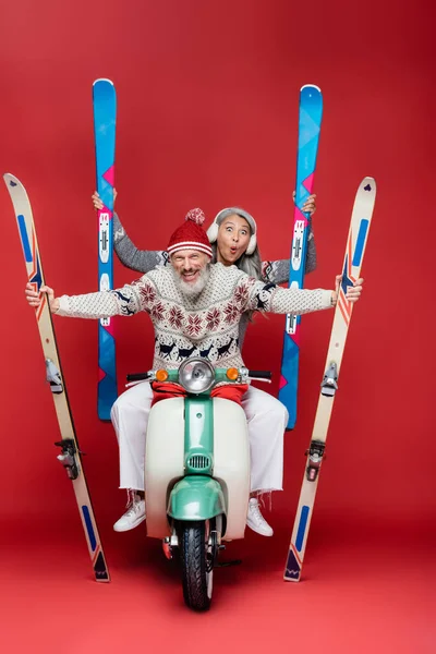 Alegre interracial e meia idade casal em camisolas segurando esquis enquanto equitação motor scooter no vermelho — Fotografia de Stock
